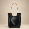 Black woven tote bag with long tan handles on a warm ivory studio background
