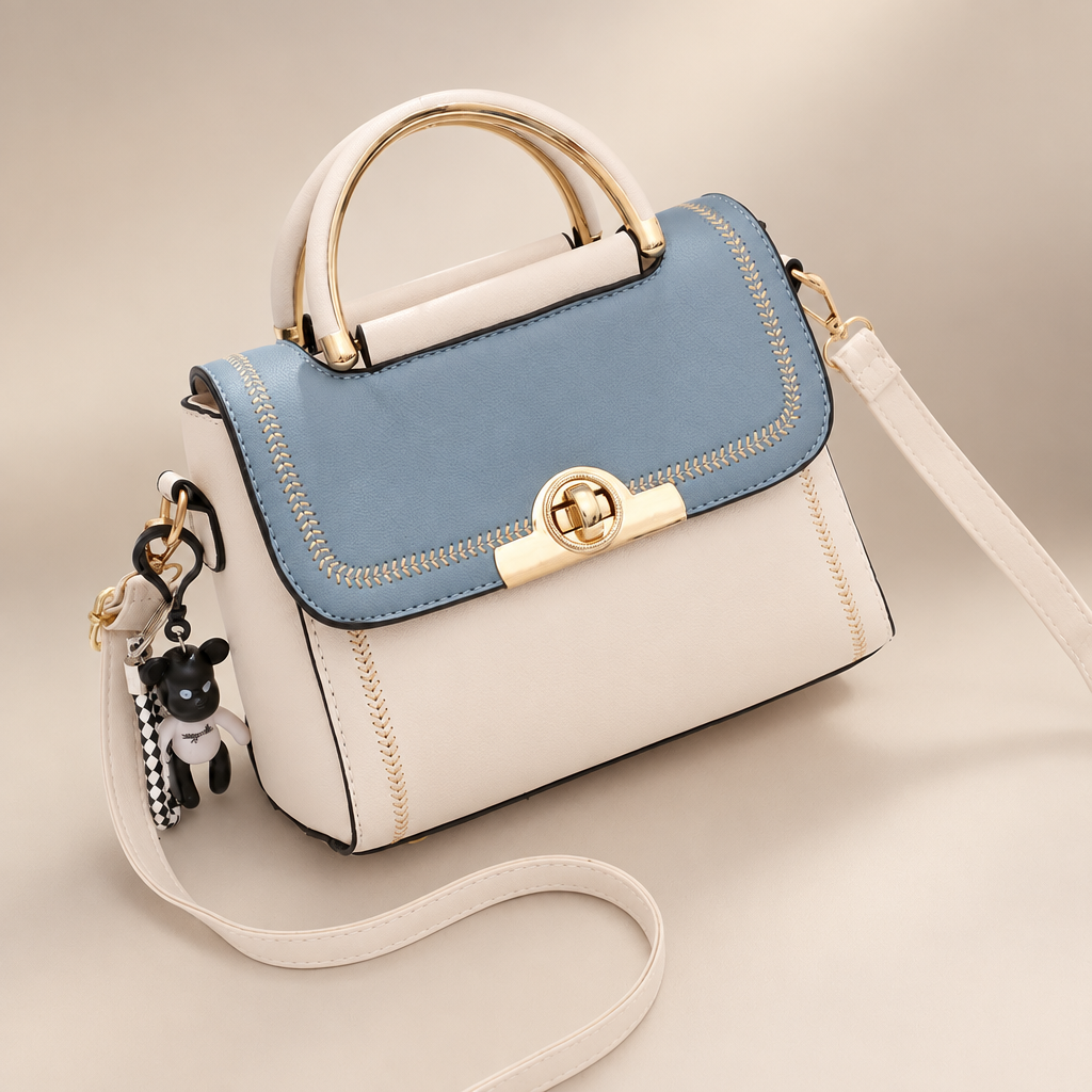 Blue And White embroidered crossbody bag with pale body, blue flap, top handle and key charm on a warm ivory and taupe studio background