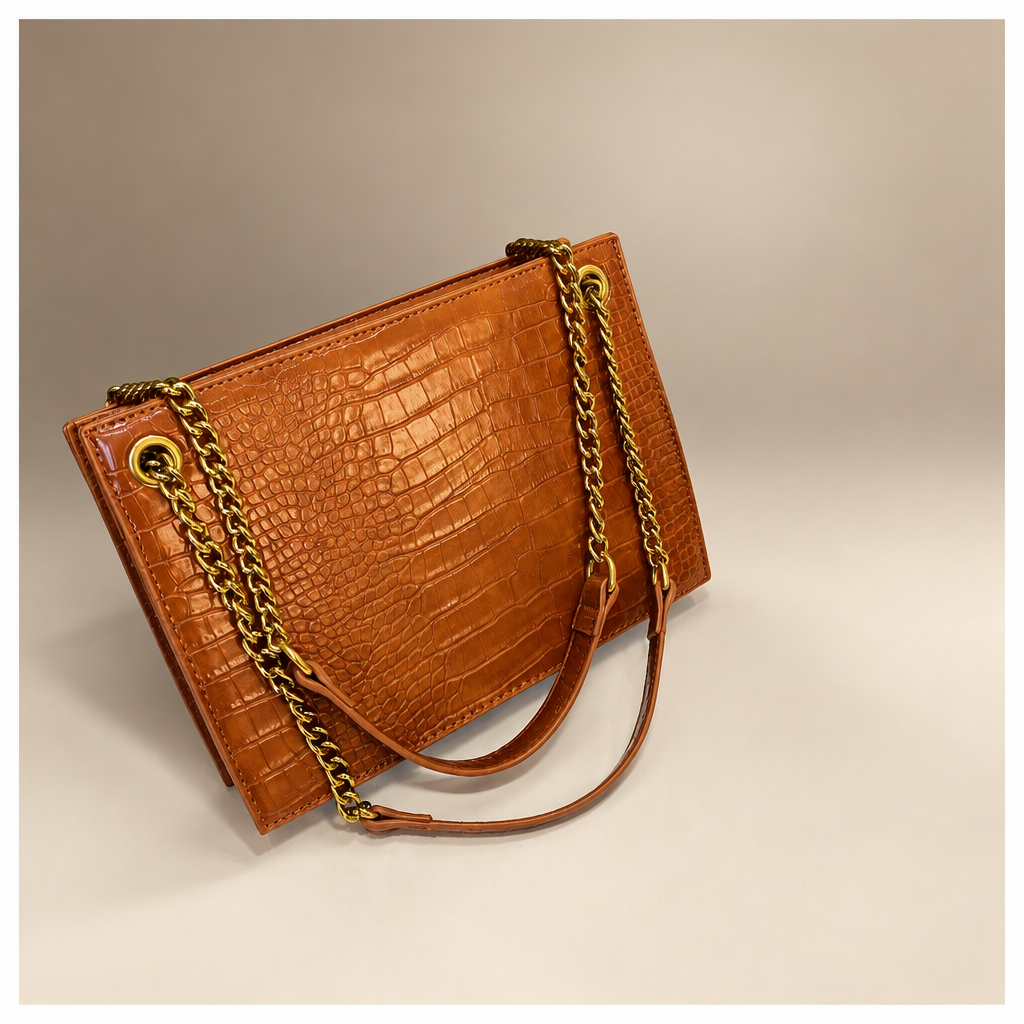 Brown crocodile-pattern shoulder bag with gold chain straps on a warm ivory and taupe studio background