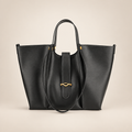 Black tote bag with gold-tone oval hardware and long strap on a warm ivory studio background